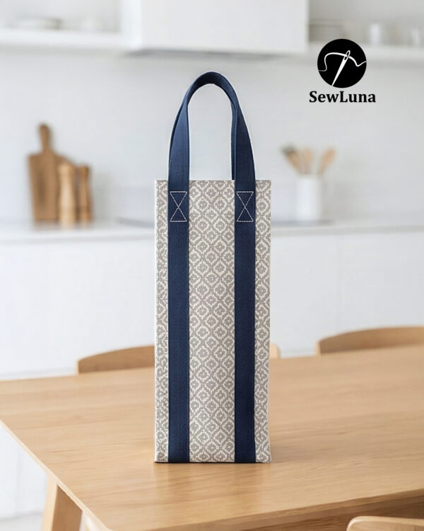 Handmade fabric wine carrier with blue straps and a grey geometric diamond pattern sitting next to a red wine bottle with a burgundy bow on a wooden table.