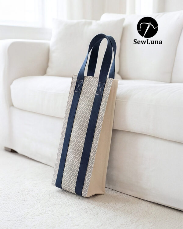 Handmade fabric wine carrier with blue straps and a grey geometric diamond pattern sitting next to a red wine bottle with a burgundy bow on a wooden table.