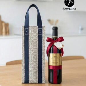 Handmade fabric wine carrier with blue straps and a grey geometric diamond pattern sitting next to a red wine bottle with a burgundy bow on a wooden table.