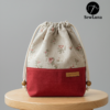 Finished example of the Bloom Essential Drawstring Pouch sewing pattern sitting on a wooden stool. The bag features a beige floral top section and a solid red canvas bottom with a leather handmade tag. It has a gathered drawstring top with cotton cord and wooden beads.