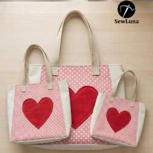 Three handmade cream and pink polka-dot canvas tote bags in small, medium, and large sizes featuring a large red heart applique on the front panel, displayed on a light wood surface.