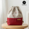Finished example of the Bloom Essential Drawstring Pouch sewing pattern sitting on a wooden stool. The bag features a beige floral top section and a solid red canvas bottom with a leather handmade tag. It has a gathered drawstring top with cotton cord and wooden beads.