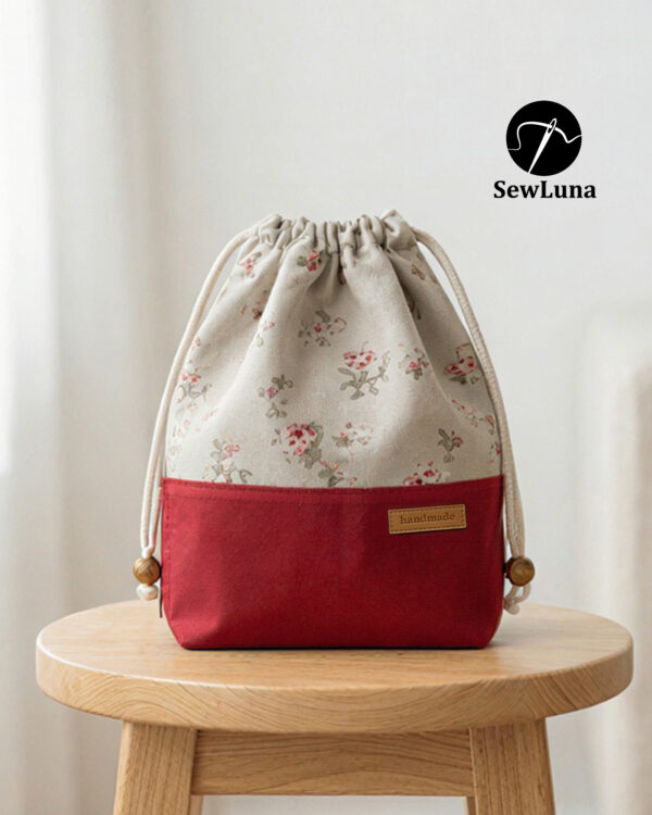 Finished example of the Bloom Essential Drawstring Pouch sewing pattern sitting on a wooden stool. The bag features a beige floral top section and a solid red canvas bottom with a leather handmade tag. It has a gathered drawstring top with cotton cord and wooden beads.