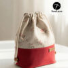 Finished example of the Bloom Essential Drawstring Pouch sewing pattern sitting on a wooden stool. The bag features a beige floral top section and a solid red canvas bottom with a leather handmade tag. It has a gathered drawstring top with cotton cord and wooden beads.