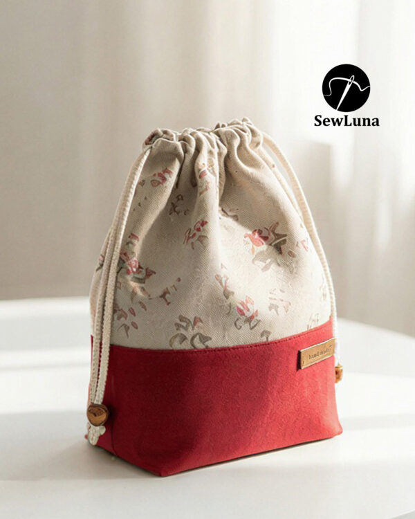 Finished example of the Bloom Essential Drawstring Pouch sewing pattern sitting on a wooden stool. The bag features a beige floral top section and a solid red canvas bottom with a leather handmade tag. It has a gathered drawstring top with cotton cord and wooden beads.