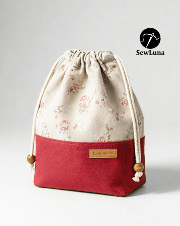 Finished example of the Bloom Essential Drawstring Pouch sewing pattern sitting on a wooden stool. The bag features a beige floral top section and a solid red canvas bottom with a leather handmade tag. It has a gathered drawstring top with cotton cord and wooden beads.