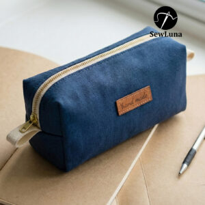 Finished dark blue linen boxy zipper pouch with a tan leather handmade label and cream zipper tape, sitting on brown notebooks next to a pen. This image represents the finished result of the Finn Utility Pouch sewing pattern.