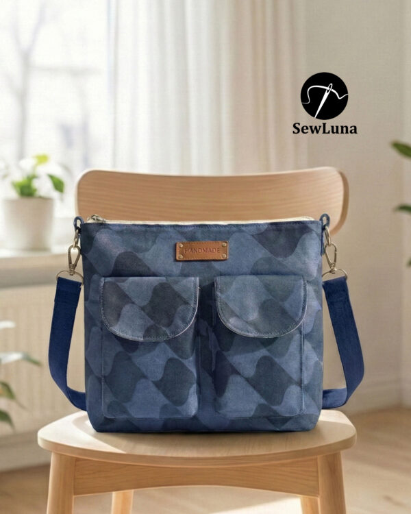 Blue patterned handmade canvas crossbody bag sitting on a wooden table near a window. This beginner-friendly PDF sewing pattern bag features a top zipper, adjustable navy strap, and two front pockets with a brown leather handmade tag.