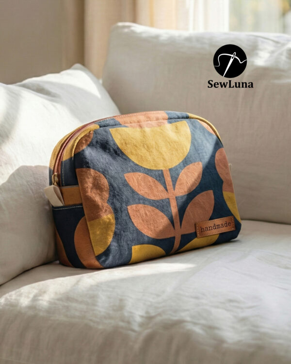 Two handmade curved zipper pouches in mustard yellow and navy blue botanical linen fabric sitting on a rustic wooden table. The larger toiletry bag stands behind a smaller matching cosmetic case which features a brown leather "handmade" label.
