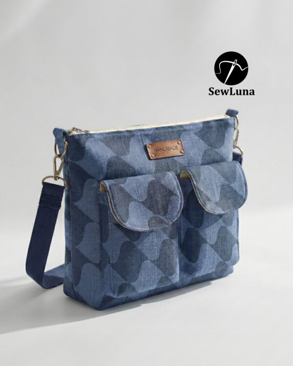 Blue patterned handmade canvas crossbody bag sitting on a wooden table near a window. This beginner-friendly PDF sewing pattern bag features a top zipper, adjustable navy strap, and two front pockets with a brown leather handmade tag.