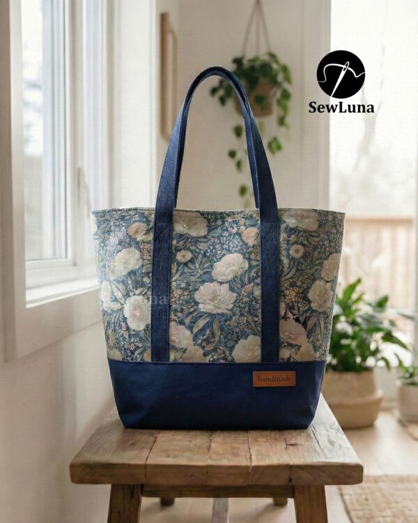 Finished Lyra canvas tote bag sewing pattern featuring a vintage blue floral tapestry upper, solid navy blue base, and navy webbing straps. The handmade bag is standing upright against a white background, showcasing this beginner DIY project.