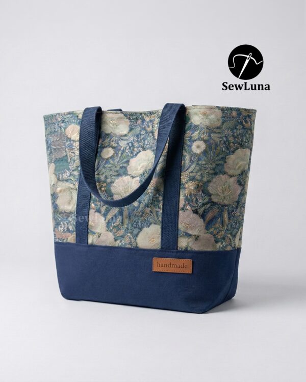 Finished Lyra canvas tote bag sewing pattern featuring a vintage blue floral tapestry upper, solid navy blue base, and navy webbing straps. The handmade bag is standing upright against a white background, showcasing this beginner DIY project.