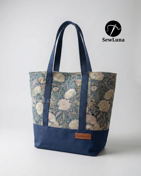 Finished Lyra canvas tote bag sewing pattern featuring a vintage blue floral tapestry upper, solid navy blue base, and navy webbing straps. The handmade bag is standing upright against a white background, showcasing this beginner DIY project.
